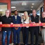 Group of eight people at a ribbon-cutting ceremony, holding a red 'ipromoteU' ribbon across the doorway.