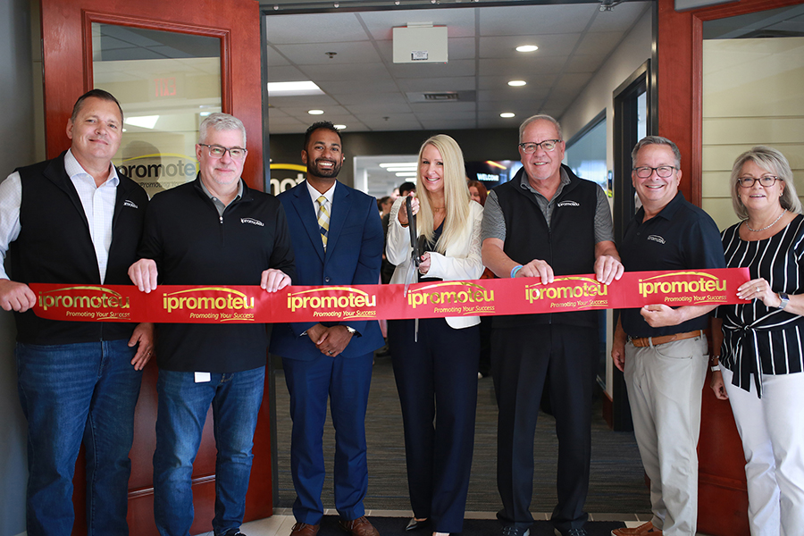Group of eight people at a ribbon-cutting ceremony, holding a red 'ipromoteU' ribbon across the doorway.
