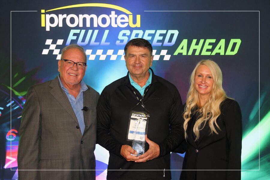 Three adults pose at a racing-themed event; the center man holds a trophy, with a bright backdrop reading 'ipromoteu' and 'FULL SPEED AHEAD'.