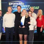 Group of eight on stage; central woman in a black blazer holds a blue trophy, smiling with colleagues around her, backdrop reads 'PromoteU' and 'FUN SPEED AHEAD'.] ,
