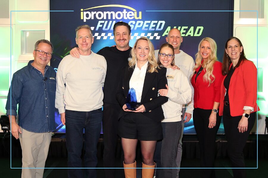 Group of eight on stage; central woman in a black blazer holds a blue trophy, smiling with colleagues around her, backdrop reads 'PromoteU' and 'FUN SPEED AHEAD'.] ,